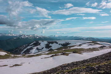 View from sunlit snowfield on hill top to snow cornice on rocky ridge against big snowbound mountain range far away under dramatic cloudy sky. Sunlight on snowy field in high mountains under clouds.
