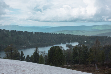 Bleak view to long mountain lake with little house in dense taiga under gray dramatic cloudy sky during heavy rain. Small country house on shore of alpine lake in thick forest in gloomy rainy weather.