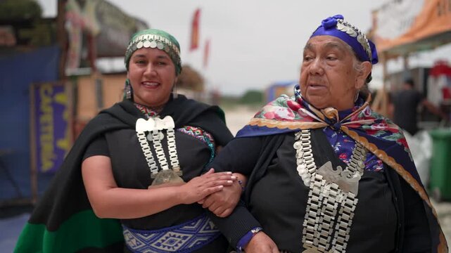 Two Mapuche women in traditional clothing and silver jewelry walking and talking at a cultural fair
