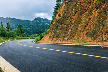 Scenic winding asphalt road curving around a rocky mountain in a rural landscape under a cloudy sky.