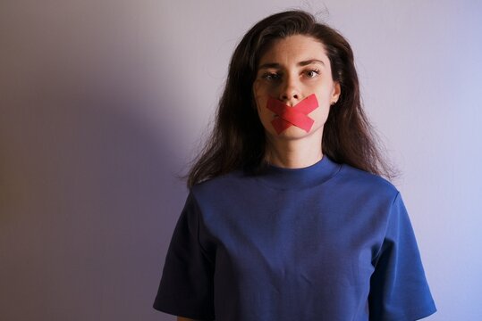 Young European brunette woman in purple t-shirt with red tape X over mouth on studio background. Concept of silencing women, feminism, protest, rights