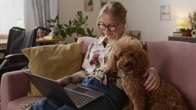 Joyful Caucasian young woman with paraplegia stroking cute miniature poodle while having video call on laptop at home
