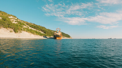 A cargo ship rests aground on a rocky shore, surrounded by trees and hills under a clear blue sky. The calm water reflects the sunlight. An abandoned ship has run aground.