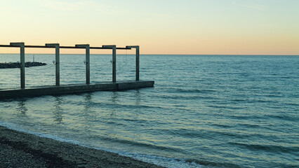 A serene view of the calm ocean at sunset, featuring a partially constructed pier with concrete pillars. Gentle waves lap against the shore, reflecting the soft pastel sky.
