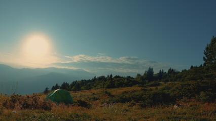 A vibrant green tent nestled in a mountainous landscape under a bright sun. The scene features lush, wild grass and distant, hazy mountains, evoking a serene camping atmosphere.