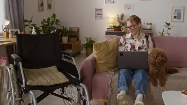 Portrait of Caucasian cheerful young woman with paraparesis posing for camera sitting on couch with laptop next to small brown dog