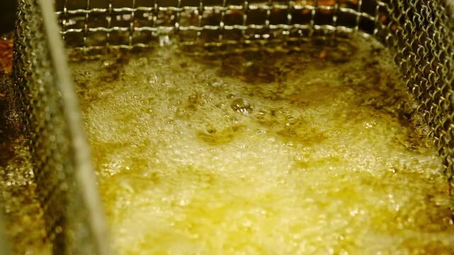Stainless strainer with potato slices in boiling oil of fryer closeup. Crispy French fries preparation in restaurant kitchen. Fast food snacks