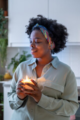 Dreamy smiling black woman holding lighted scented candle in hands with eyes closed feels comfort....