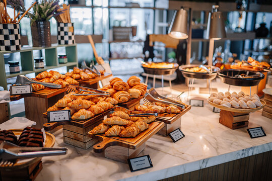 Assorted pastries including croissants and buns displayed on a marble countertop in a modern cafe with warm lighting and wooden accents during breakfast service