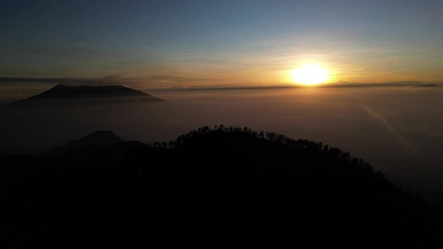 Stunning sunset aerial footage capturing the majestic silhouette of the Arjuno Welirang mountain range with mist in the valleys. Ideal for dramatic travel, nature, and Indonesian landscape projects.