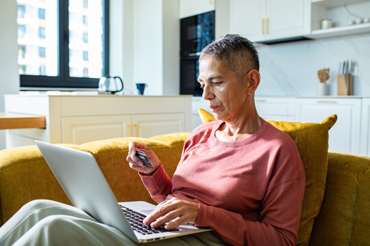 Older woman using laptop and credit card on sofa at home