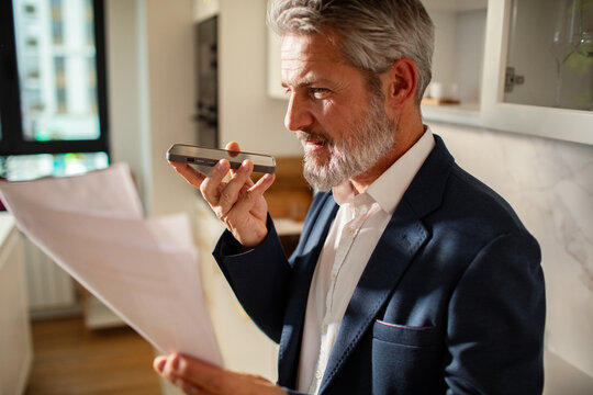Mature businessman dictating on smartphone while reviewing documents at home