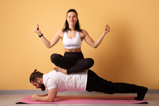 Fit couple practicing unique yoga balance pose, woman seated on man in plank position on pink mat, warm studio background, active lifestyle concept.