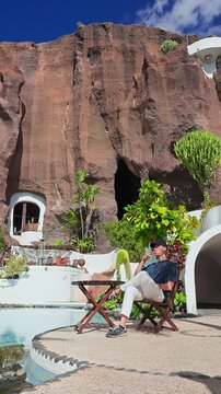 Sunlit Lagomar Museum courtyard in Lanzarote shows a man seated by a curved pool, sipping a drink near cacti and succulents, with cave, white forms, and strong midday light.