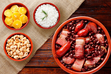 A complete Brazilian feijoada served in a clay pot with black beans, sausage, bacon, and pork, accompanied by white rice, orange slices, farofa and (pork crackling) on rustic wooden table