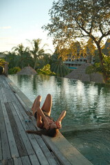 Young woman in black swimsuit relaxing and lying on the edge of luxury infinity pool during sunset at tropical resort.