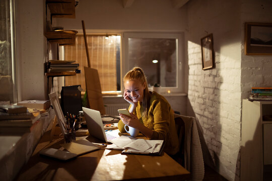 Young woman using smartphone and laptop in cozy home office at night