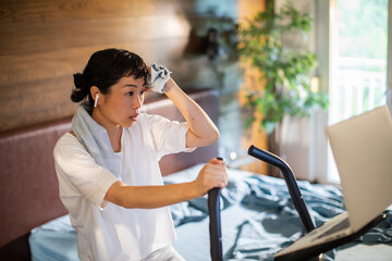 Woman cycling on exercise bike in bedroom with laptop