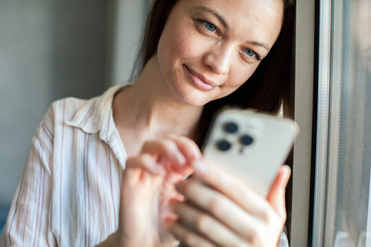 Smiling woman using smartphone by window at home