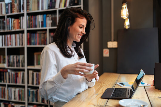 Side view of a woman in a white shirt frinking coffee while using a laptop at a wooden cafe table with coffee, warm hanging lights and bookshelves in the background for study or remote work.