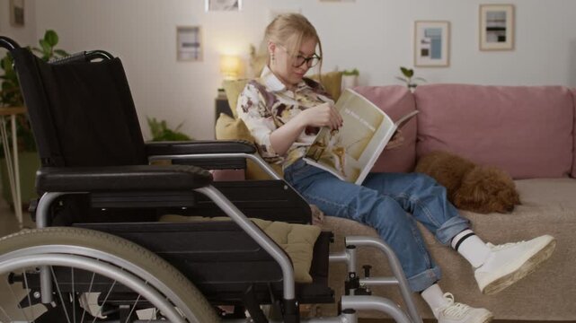 Rack focus shot of wheelchair and gen Z girl with disability looking through magazine while relaxing on couch next to brown dog