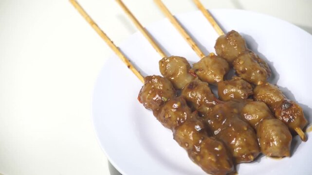 Grilled meatballs or Bakso Bakar on skewers with spicy sweet soy sauce. Popular Indonesian street food snack, smoky BBQ beef balls, spicy condiment, rotating on white plate, delicious meat appetizer, 
