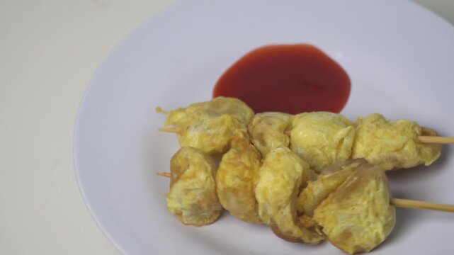 Fried egg-wrapped meatballs or Cilor snack on skewers with chili sauce. Traditional Indonesian street food, savory egg roll snack, rotating on white plate, popular school snack, crispy omelet texture,