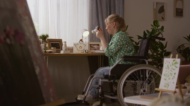 Blonde gen Z girl with disability taking photos of decorative items placed on her table while sitting in wheelchair and practicing photography with new camera