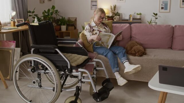 Blonde girl with disability sitting on couch with her pet and reading magazine in living room, with black wheelchair in foreground