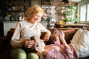 Grandmother and granddaughter with hearing aids watching smartphone on sofa at home
