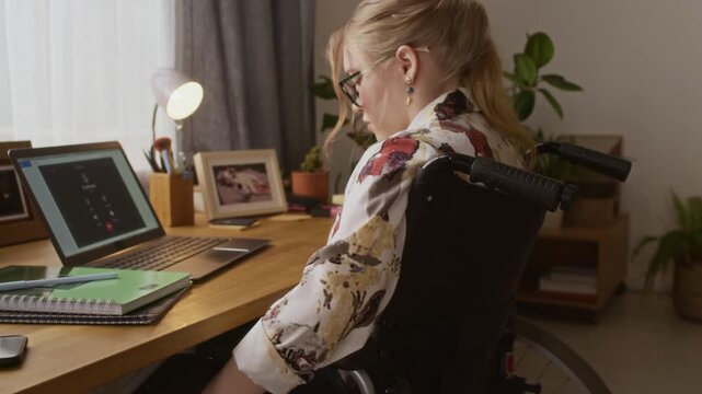 Tilt up view of Caucasian young woman in wheelchair pushing herself to wooden desk with notebooks and laptop displaying video call application