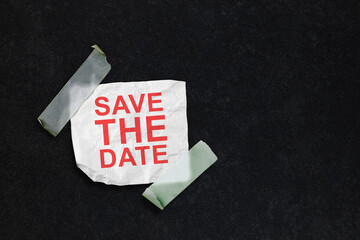 A conceptual image featuring the text 'Save the Date' in red on a crumpled piece of white paper, taped to a dark textured background with translucent tape. Great for event reminders