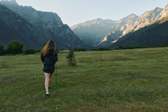 A lone hiker walks across a wide grassy meadow toward distant jagged mountains, while warm sunlight highlights the flora and creates a tranquil, expansive landscape. - Powered by Adobe