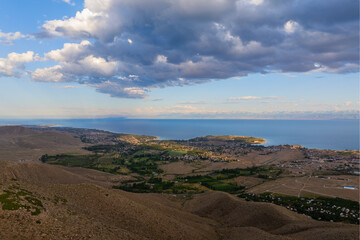 Tranquil Issyk-Kul lake reflects majestic mountains under cloudy sky with Cholpon-Ata town on the shore.
