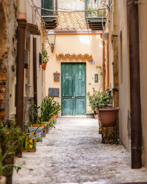 View of a quaint alleyway with vibrant green doors and potted plants lining the cobblestone path between weathered buildings, Cefalu, Sicily, Italy.