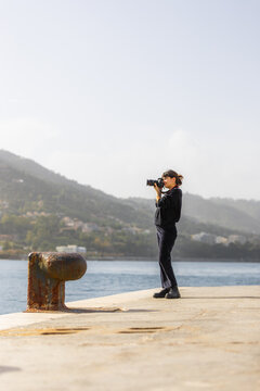 View of a photographer silhouetted against the shimmering sea, capturing the distant mountains under a pale sky, standing on a weathered concrete pier, Cefalu, Sicily, Italy.