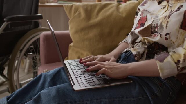 Positive girl with disability wearing glasses working on laptop and stroking small brown dog sitting down on couch next to her