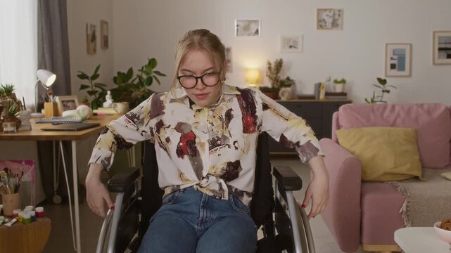 Caucasian young woman with paraparesis sitting in wheelchair pushing herself forward towards camera while moving around living room