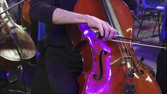 Male musician deeply engaged in playing large cello with bow moving across strings. Orchestral performance under vibrant purple stage light