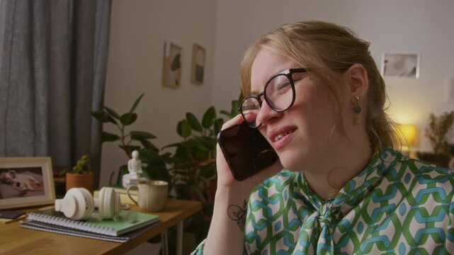 Tilt up shot of positive gen Z girl with paraplegia wearing glasses, blouse and jeans talking on phone while sitting in wheelchair next wooden desk at home