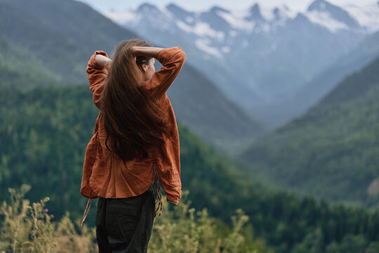 Woman stands outdoors with back to camera in a brown jacket, gazing at expansive mountains and lush valley scenery, creating a sense of freedom and peaceful adventure in nature.