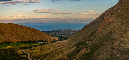 Winding road leads through peaceful mountains, overlooking a small town Cholpon-Ata by a Issyk-Kul lake. Late afternoon light enhances the majestic view.