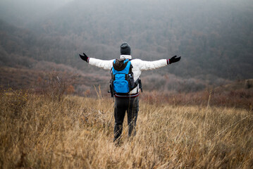 Obraz premium Rear view of hiker in warm clothing standing in nature with backpack and arms outstretched, enjoying freedom and achievement in a foggy winter landscape.