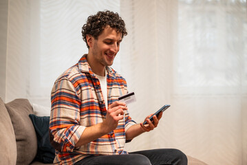 Happy young man sitting on sofa in living room, comfortably using his mobile phone for payment with credit card. Wireless technology and online shopping concept.