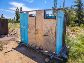 Weathered outdoor toilet structure features made with wooden doors and junk. Set in a rural, arid landscape surrounded by sparse vegetation and dirt ground.