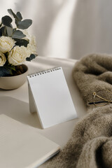 A blank white card stands on a soft surface surrounded by a cozy knit, flowers, and a book, creating a serene environment for creative ideas