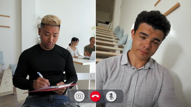 A man joins an online call but looks bored as he writes notes. He is in a room with a person seen in the background. The call takes place in daylight.