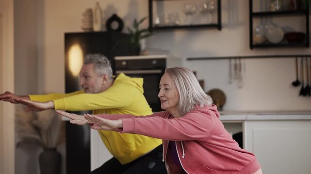 Happy middle-aged man and woman doing squats together while exercising at home