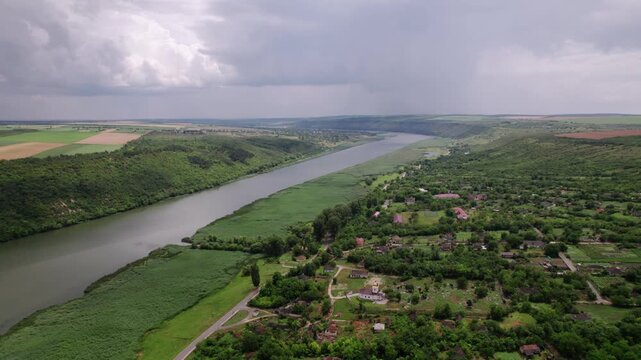 Aerial River Valley Village Under Overcast Sky, Winding River Bordering Green Banks, Scattered Rural Houses And Farm Plots, Unpaved Roads Cutting Through Pastures, Moody Cloudscape