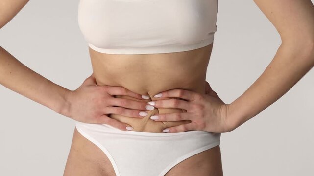 Close-up studio shot of a woman pressing her lower abdomen with both hands, wearing white underwear on a neutral background, health and pain concept.